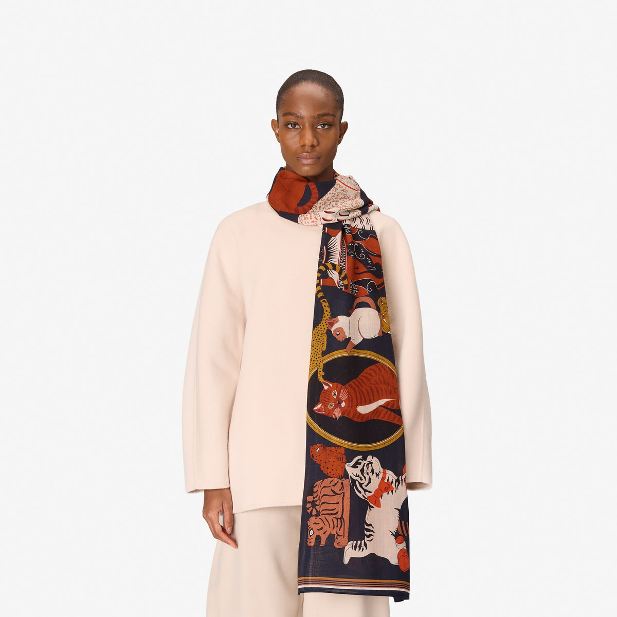 A model wearing a cream-colored coat and trousers, draped with a dark navy blue silk scarf featuring a colorful print of tigers, foxes, and a gold eagle.