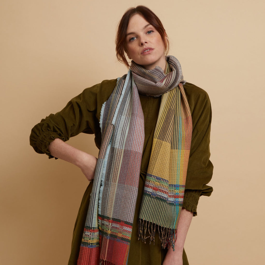 Multicolored woven wool scarf with a fringed edge, featuring a blend of grey, beige, and orange tones in a plaid pattern.