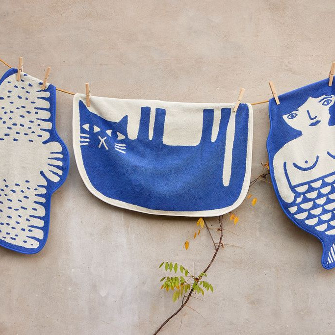 Three blue and off-white fabric cloths with animal and human illustrations hang from a string with wooden clothespins against a beige wall.
