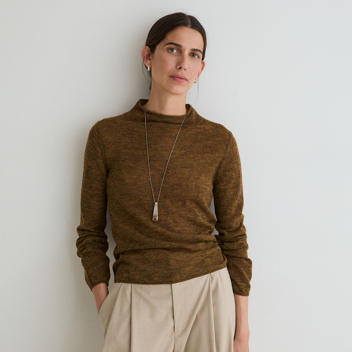 A woman wearing a brown long-sleeve knit mock neck sweater and beige wide-leg trousers, standing against a light gray wall.