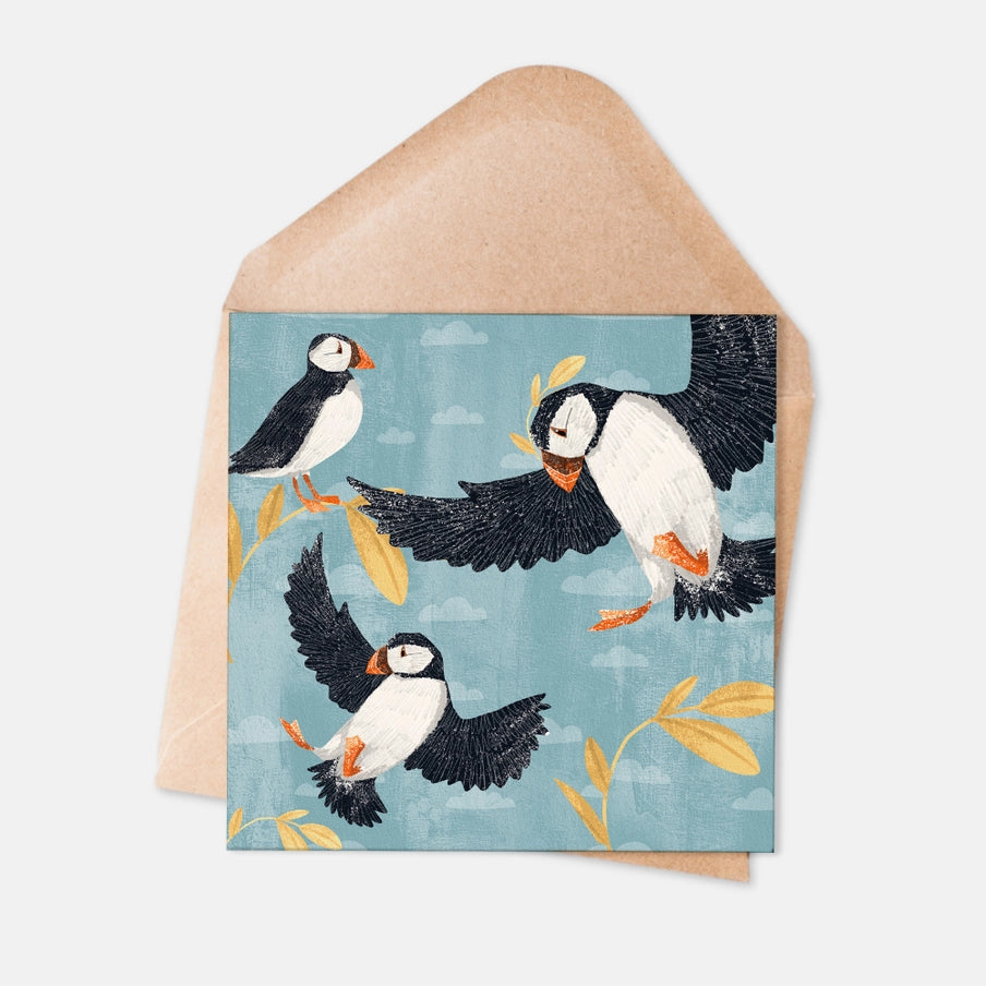 Square greeting card featuring three illustrated puffins in flight against a light blue sky with stylized foliage.