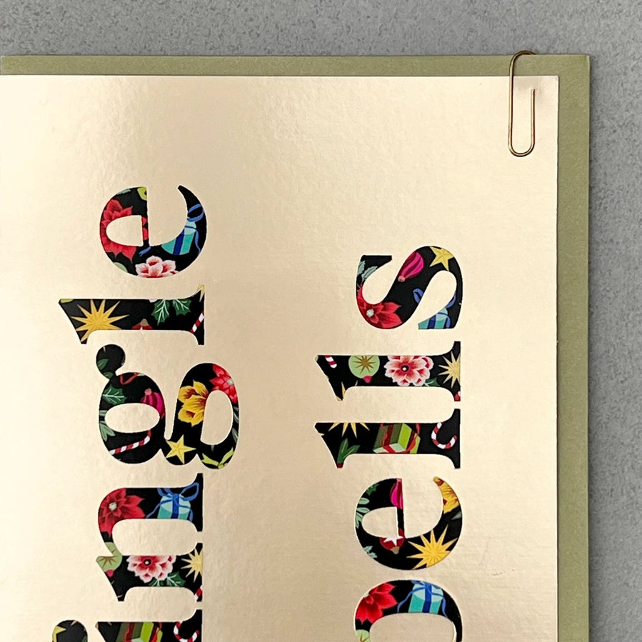 A close-up of a metallic gold foil sign featuring the word 'STUDIOS' in large letters filled with a colorful floral and geometric pattern, held by a silver paperclip against a gray textured background.