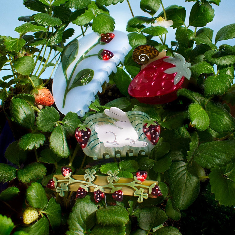 Decorative hair clips shaped like strawberries and a rabbit among green plants.