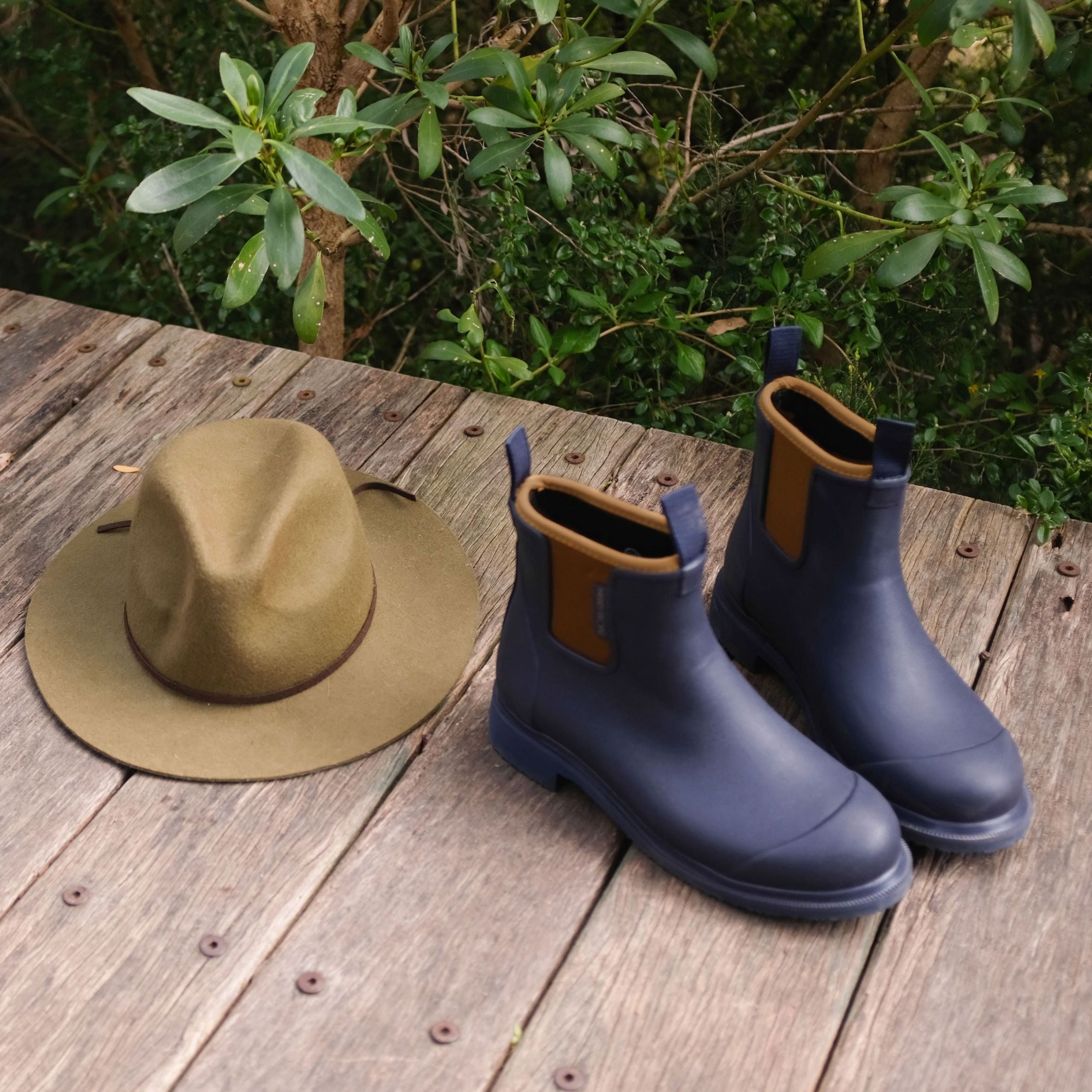 Pair of navy blue rubber rain boots with tan accents, next to a olive green felt fedora hat, on a weathered wooden surface.