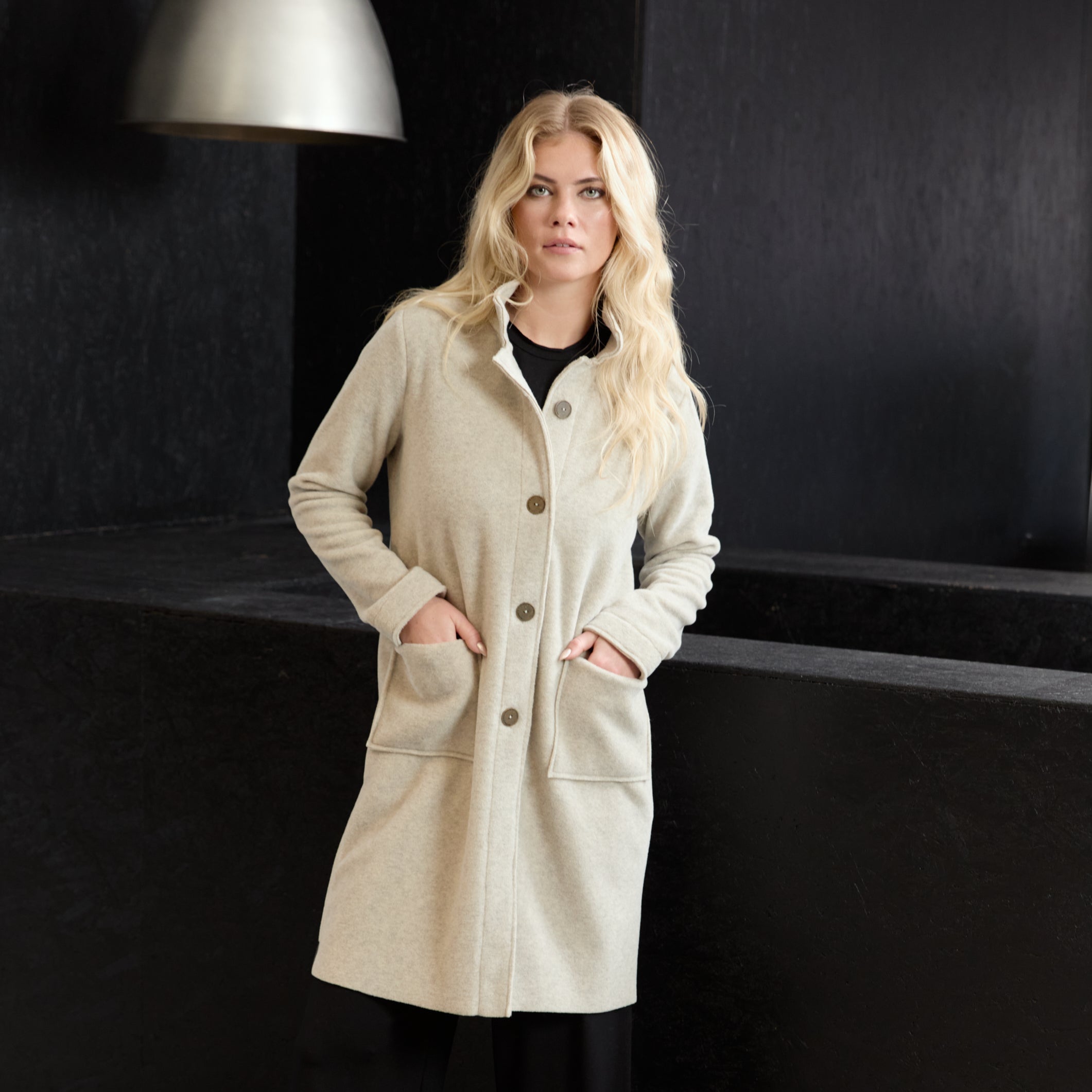 A blonde woman models a long beige wool-blend coat with front buttons and large patch pockets, paired with black wide-leg trousers and beige shoes, standing against a dark modern wall.
