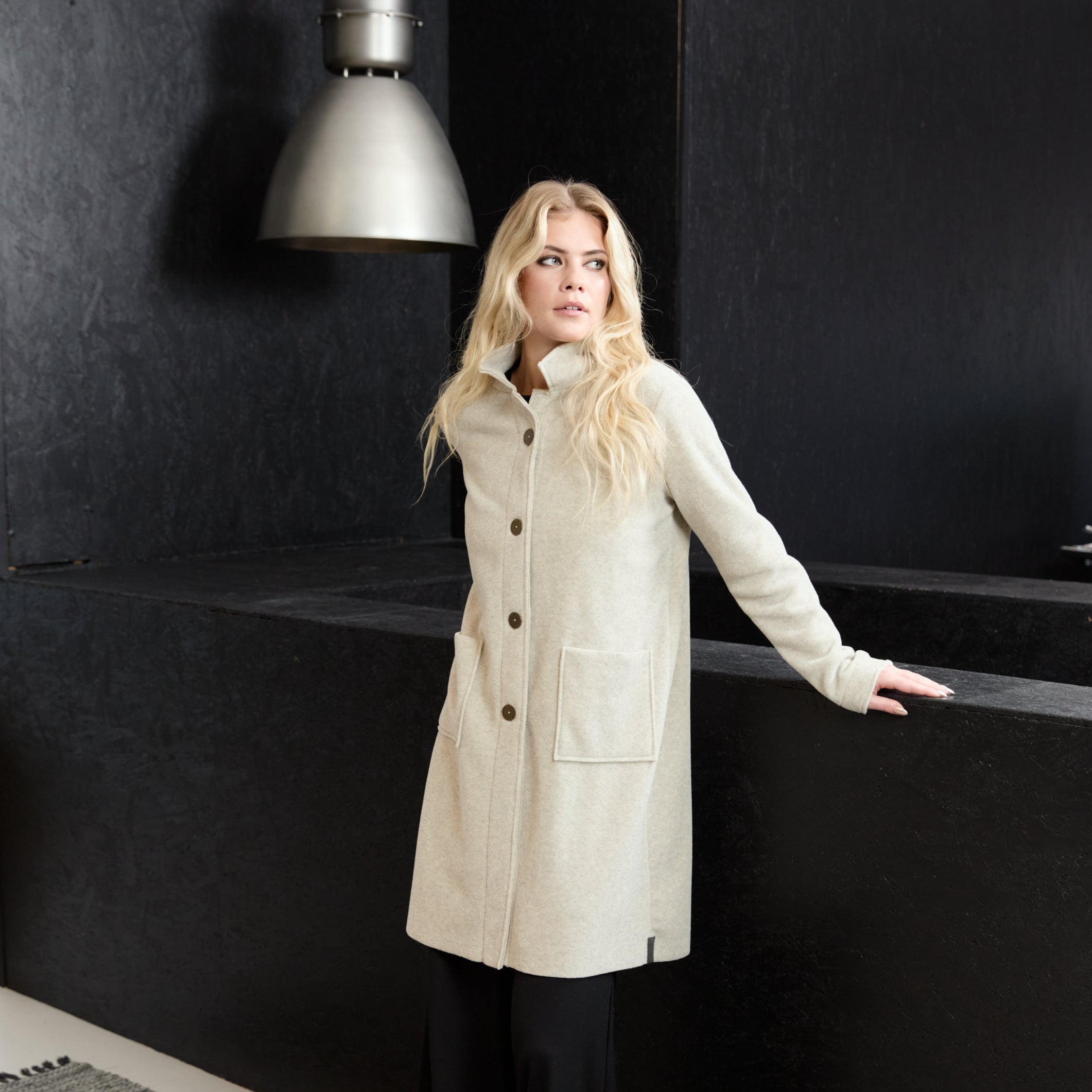 A blonde woman wearing a long cream-colored wool coat with dark buttons, black wide-leg trousers, and beige shoes, standing in a modern room with a dark textured wall and a hanging lamp.