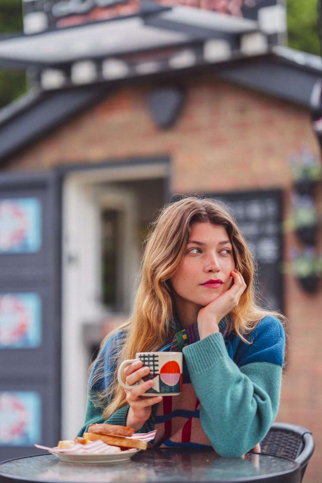 A woman with long blonde hair sits at an outdoor cafe table, holding a white ceramic mug with a colorful geometric design. She is wearing a teal and red striped knit sweater, with a blurred brick building in the background.