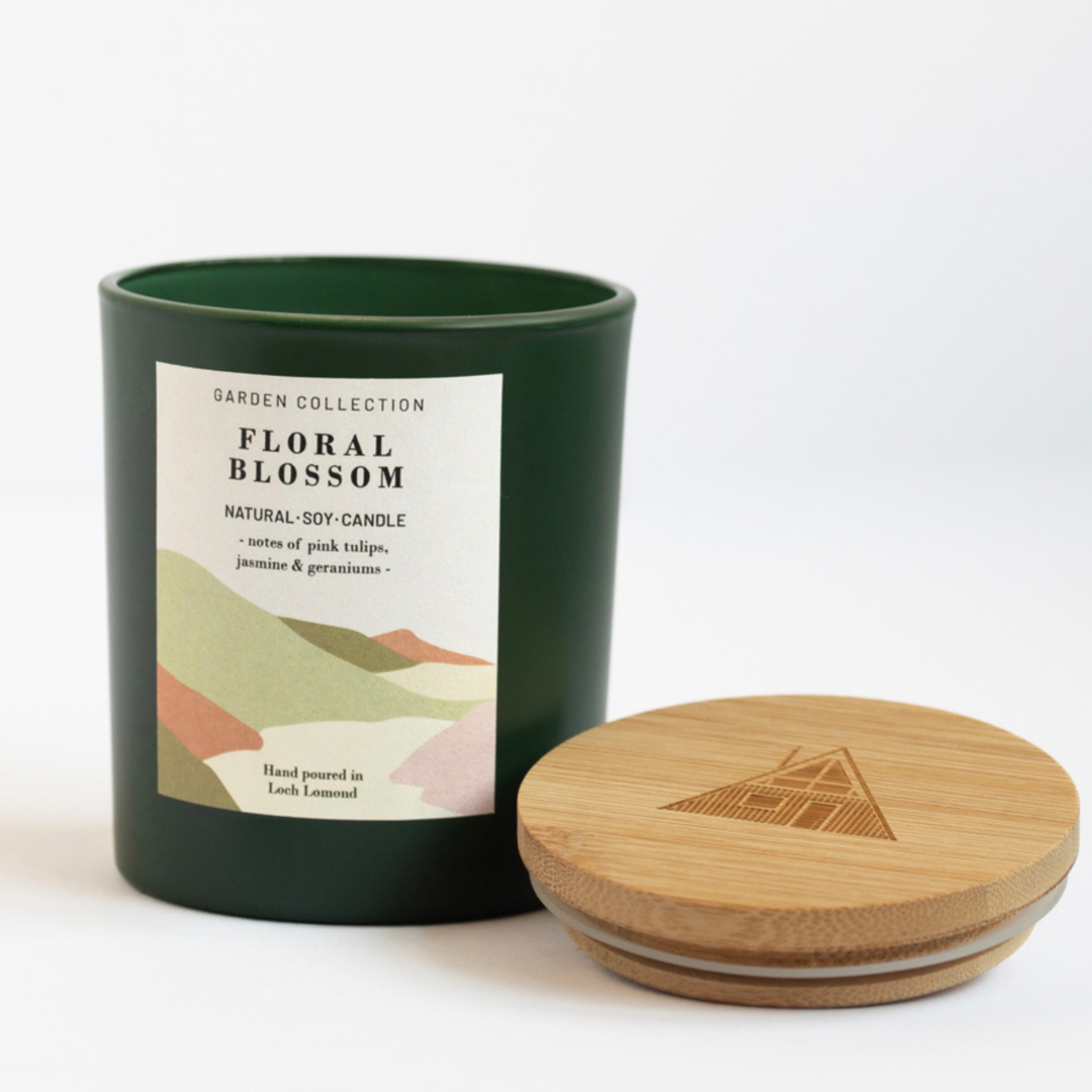 A dark green soy candle jar with a white label reading 'Garden Collection Floral Blossom' and a round natural wooden lid placed beside it on a clean white background.