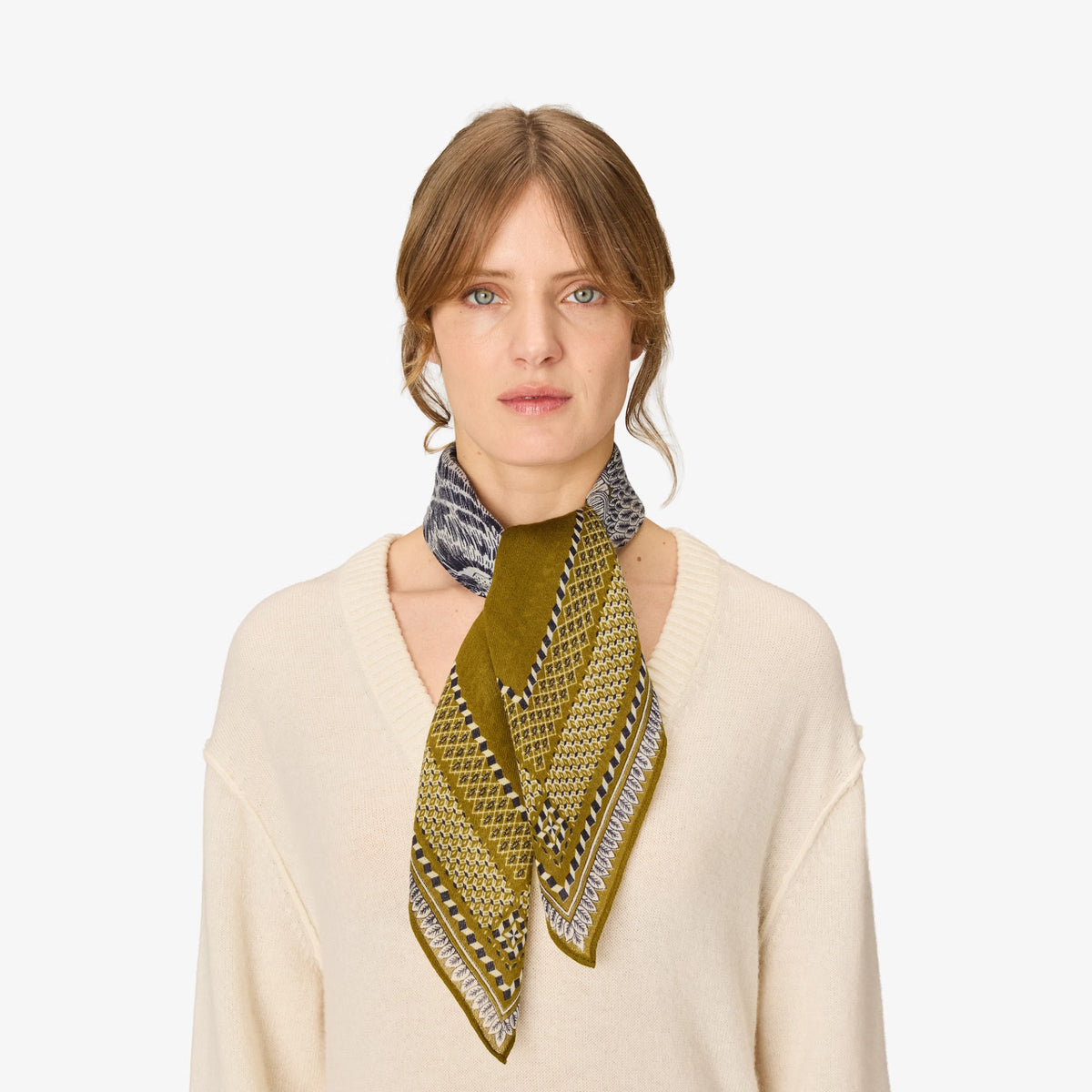 A woman wearing a cream-colored knit sweater and a patterned silk scarf tied around her neck. The scarf features a dominant olive green color with geometric patterns in black, white, and gold.