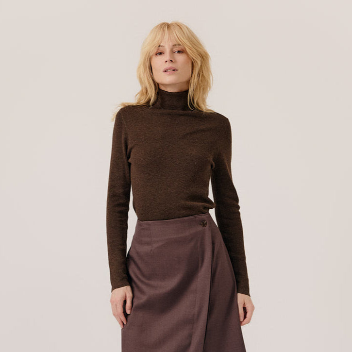 A blonde woman wearing a dark brown ribbed turtleneck sweater and a brown wrap skirt, standing against a plain light beige studio background.