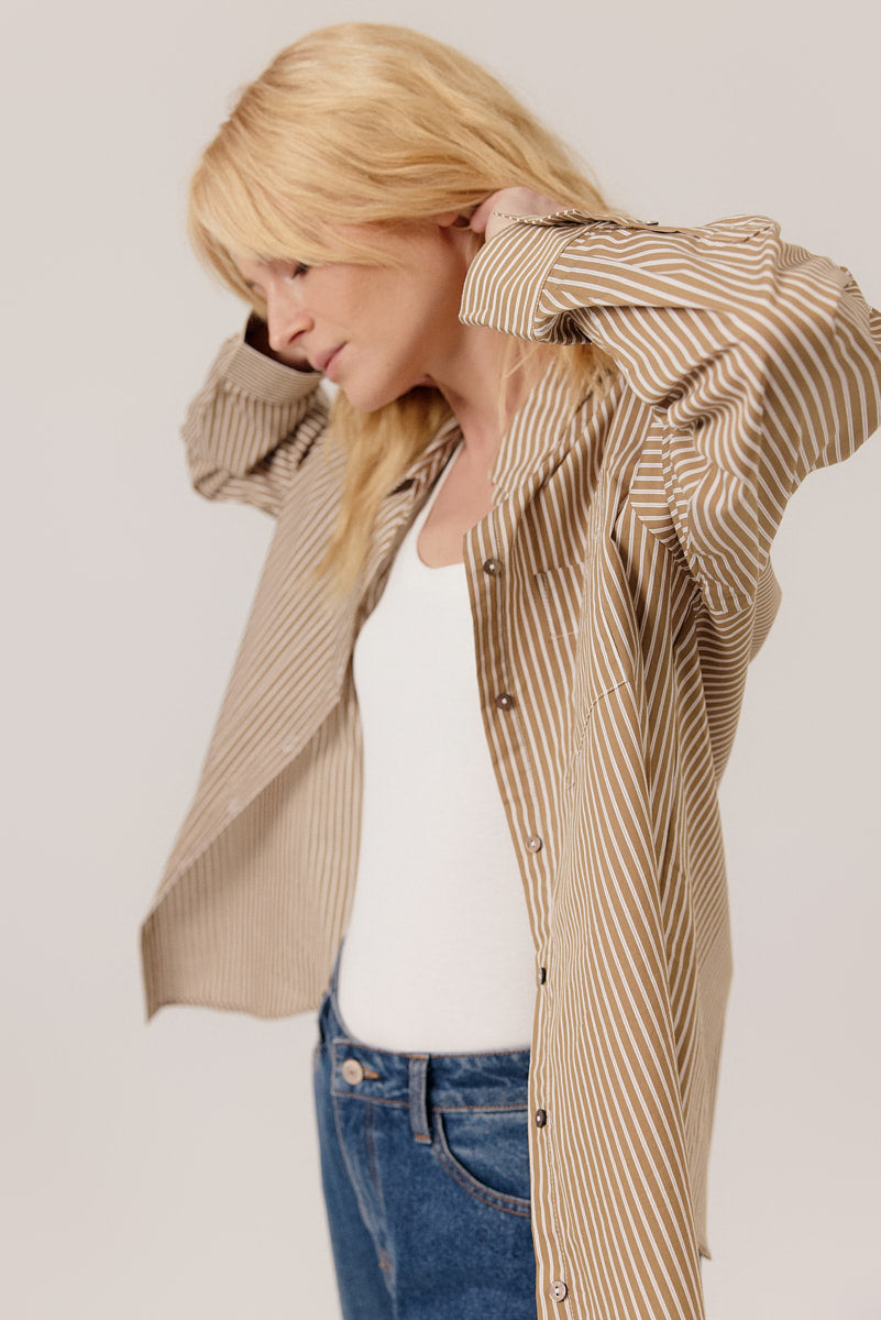 Woman wearing a beige striped shirt over a white top and blue jeans on a plain background