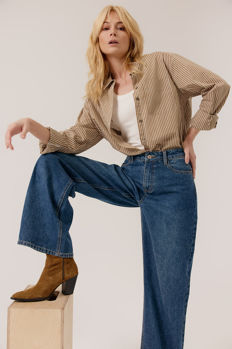 Woman wearing a striped shirt, white top, and blue jeans with brown boots on a plain background
