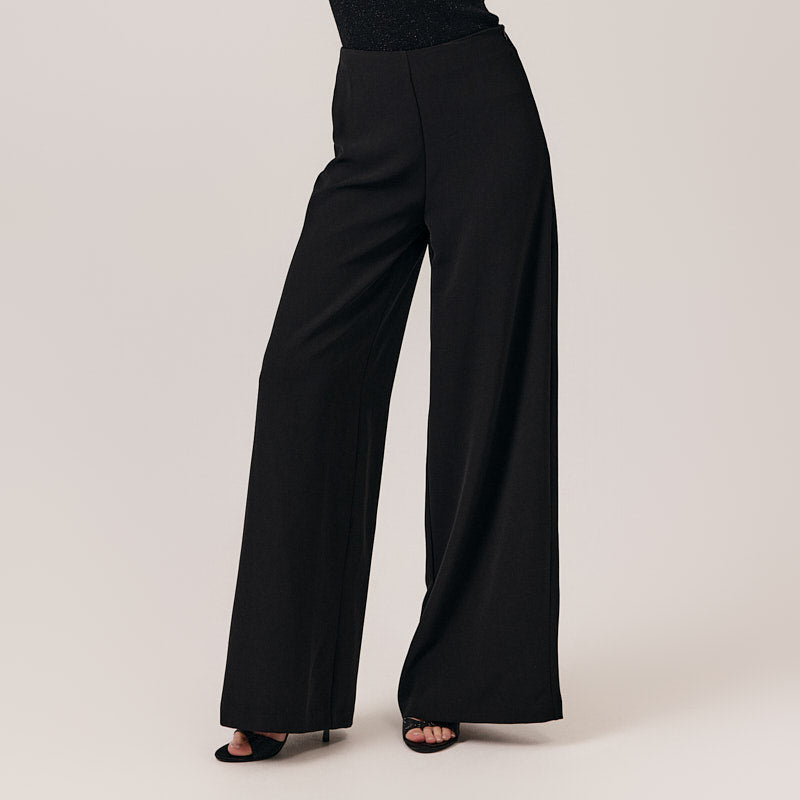 Woman wearing a black sleeveless top and wide-leg pants on a plain background