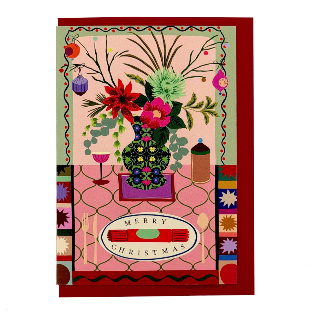 A festive Christmas greeting card featuring a colorful floral arrangement in a patterned vase, a decorated table setting, and the words 'MERRY CHRISTMAS' on a red border.