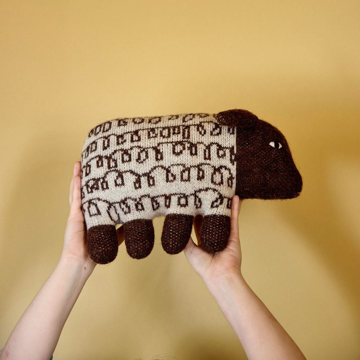 Knitted sheep toy held by hands against a yellow background
