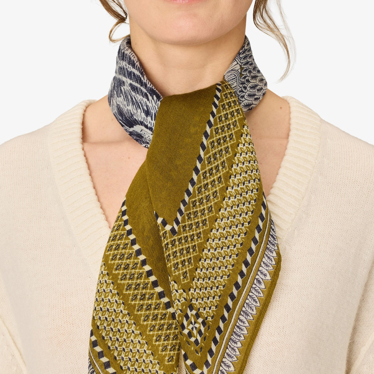 A close-up of a model wearing a patterned scarf tied around her neck over a cream-colored knit sweater. The scarf features a mix of olive green, beige, black, and white geometric and textured patterns.