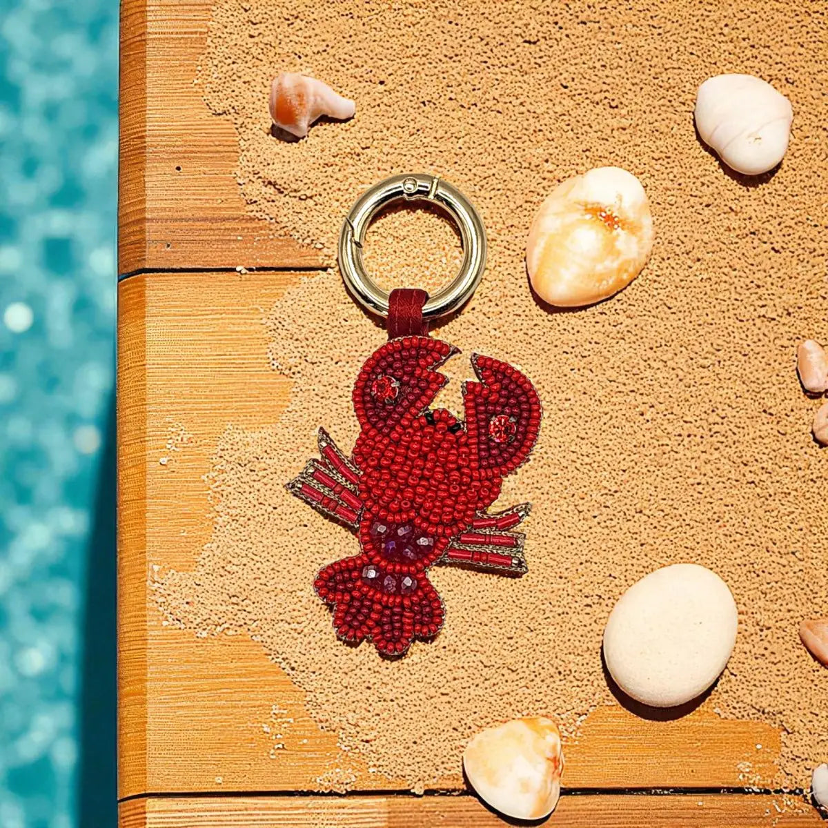 A lobster-shaped beaded keychain in deep red with small silver bead accents and a silver split ring, photographed on a wooden surface with sand and seashells nearby.