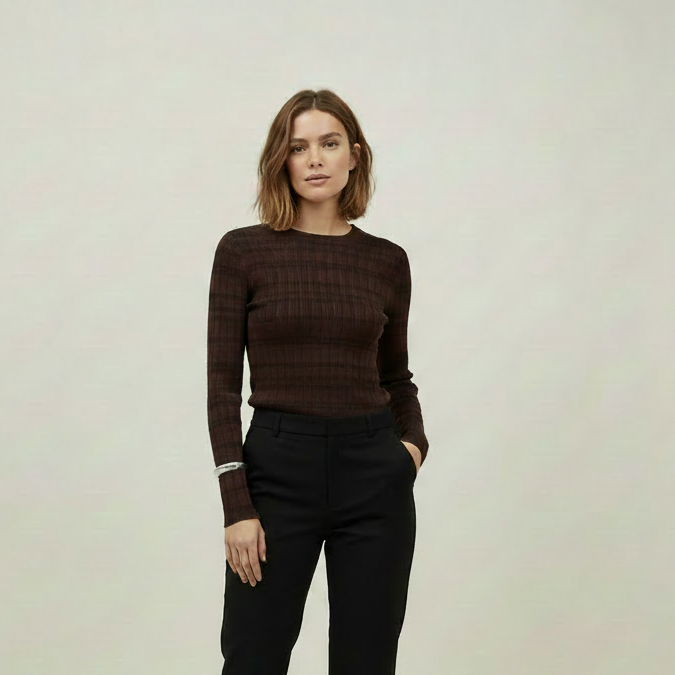 A woman stands against a plain light gray studio background wearing a dark brown textured long-sleeve top, black cropped trousers, and black-and-white patterned flats.