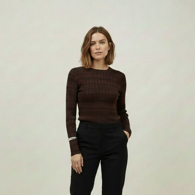A woman stands against a plain light gray studio background wearing a dark brown textured long-sleeve top, black cropped trousers, and black-and-white patterned flats.