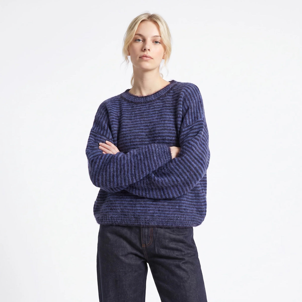 A woman modeling a dark blue and purple heathered knit crewneck sweater with dark jeans, standing against a plain white studio background.