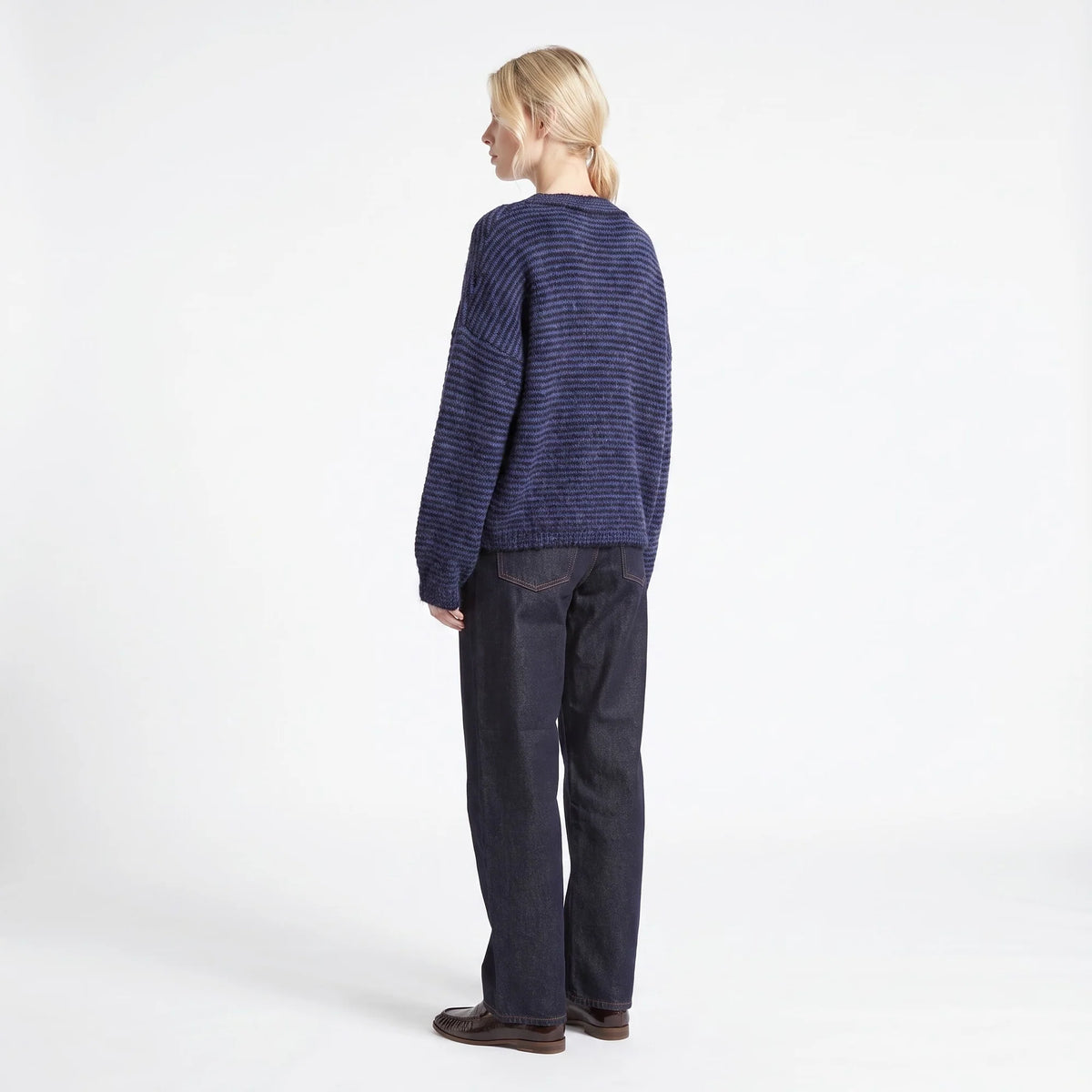 A woman stands in profile wearing a navy blue and black striped knit sweater, dark indigo jeans, and brown leather shoes against a clean white studio background.