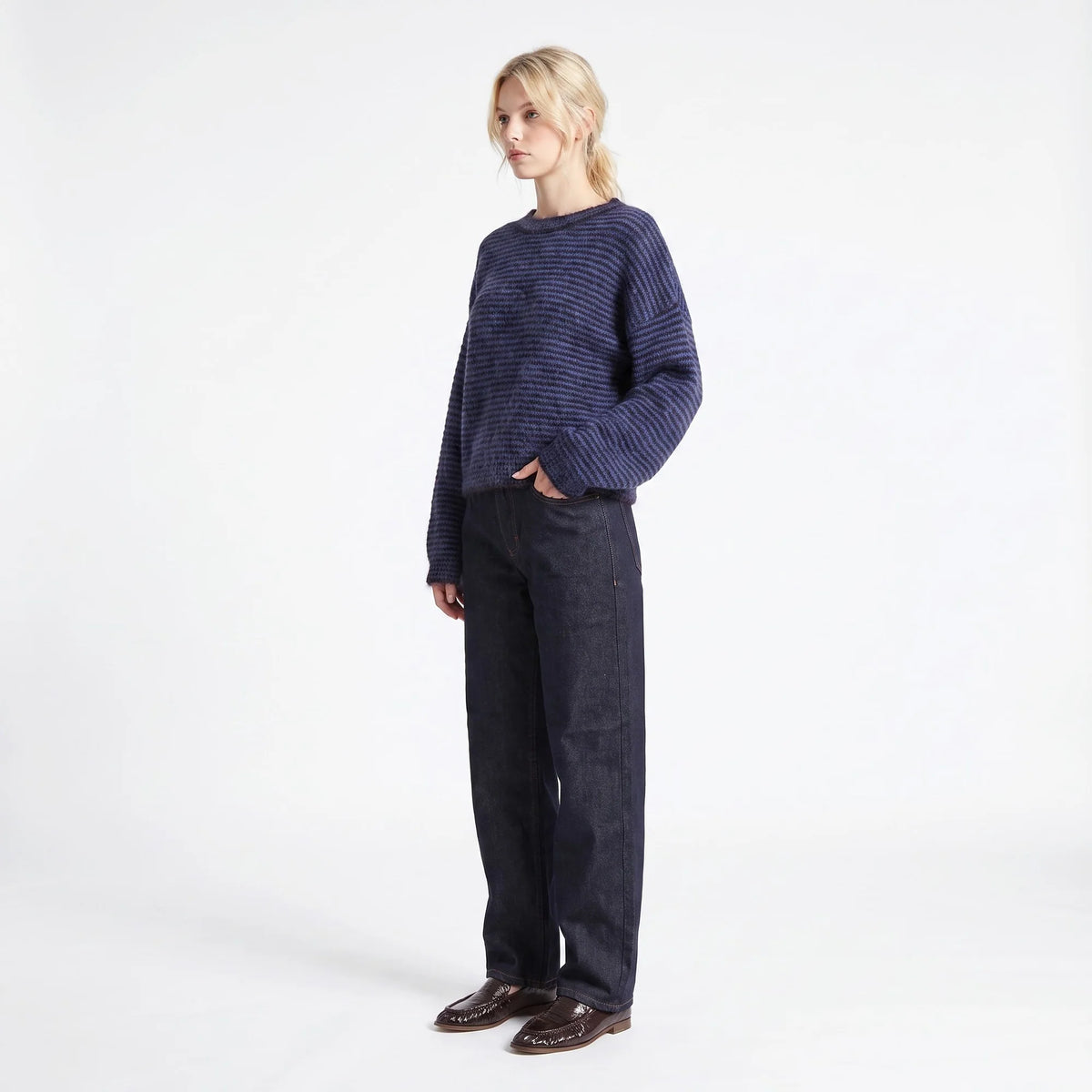 A woman stands against a plain white studio background wearing a dark navy ribbed knit sweater, dark blue jeans, and brown leather loafers.