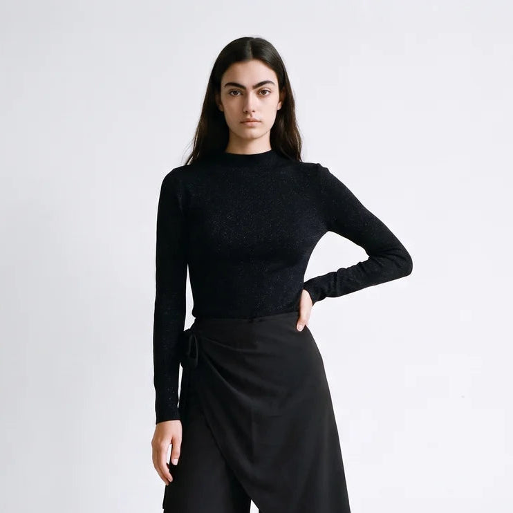 A model wearing a black long-sleeve textured mock-neck top and black wide-leg draped trousers, paired with black strappy heels, against a plain light gray studio background.
