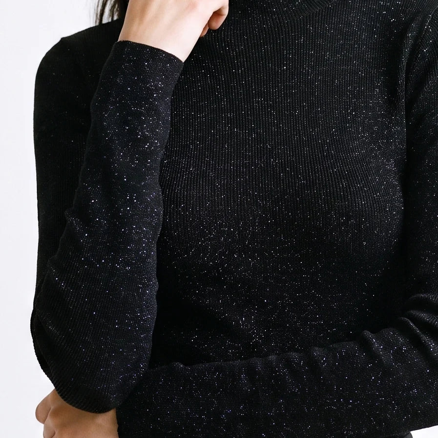 Close-up of a person wearing a black ribbed knit sweater with subtle silver glitter flecks, showing the sleeve and upper torso against a plain light background.