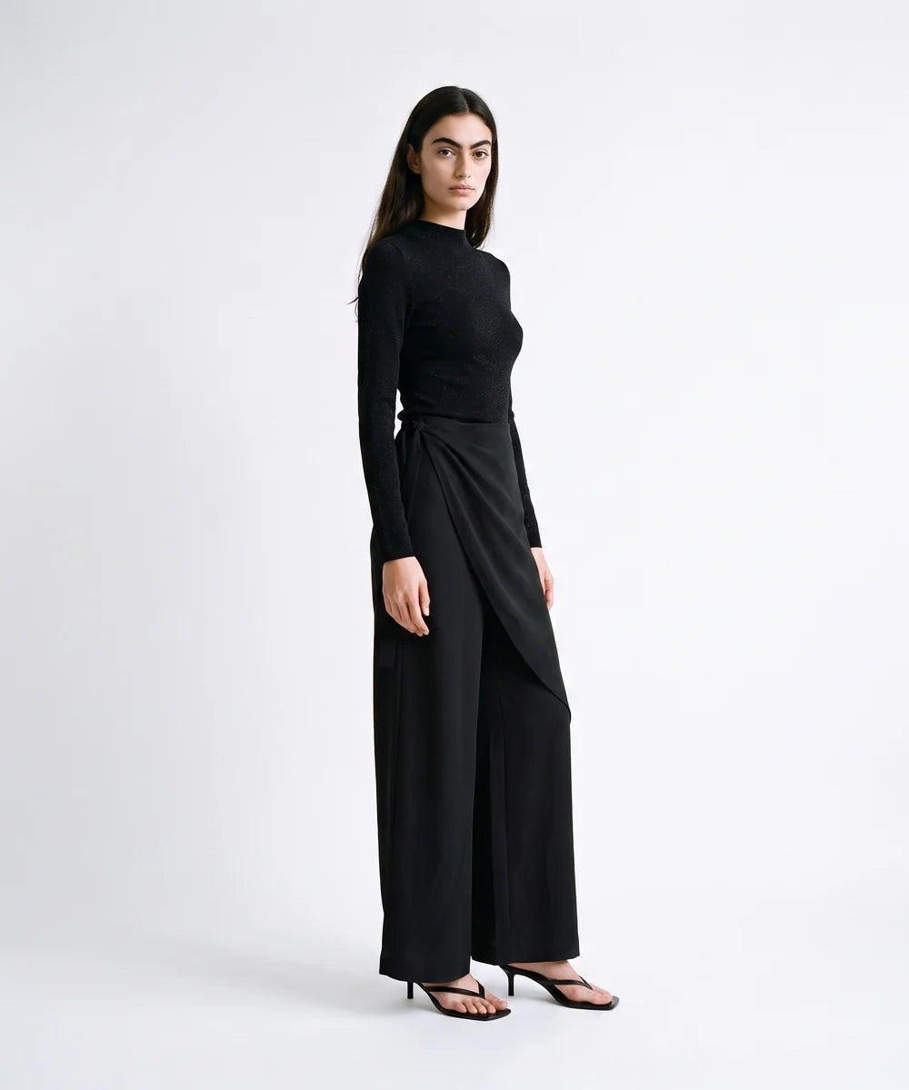 A woman modeling a black long-sleeve wrap-style top and matching wide-leg trousers, paired with black heeled sandals, against a plain light gray studio background.