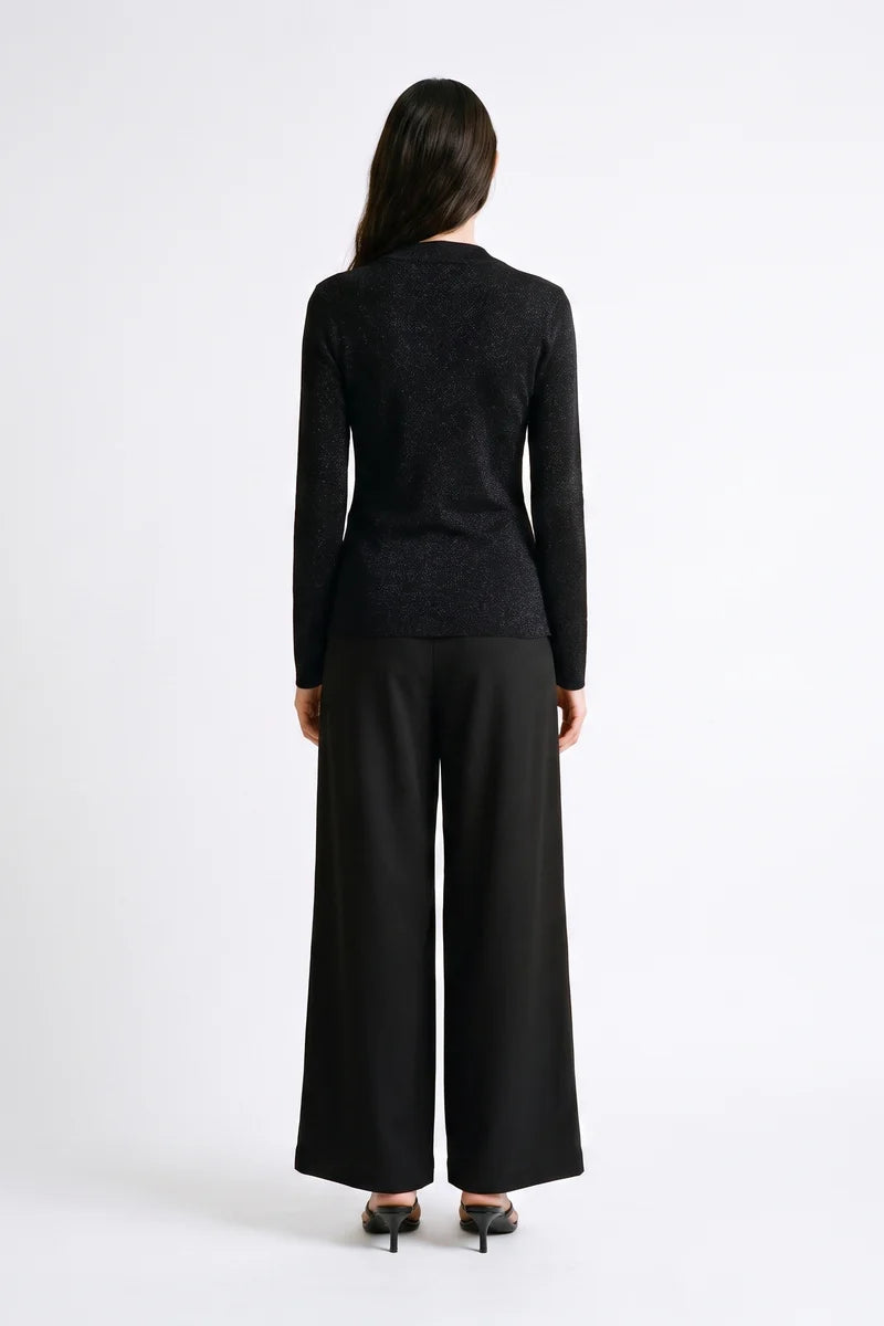 A woman viewed from the back wearing a black textured long-sleeve top and black wide-leg trousers, standing against a plain white studio background.