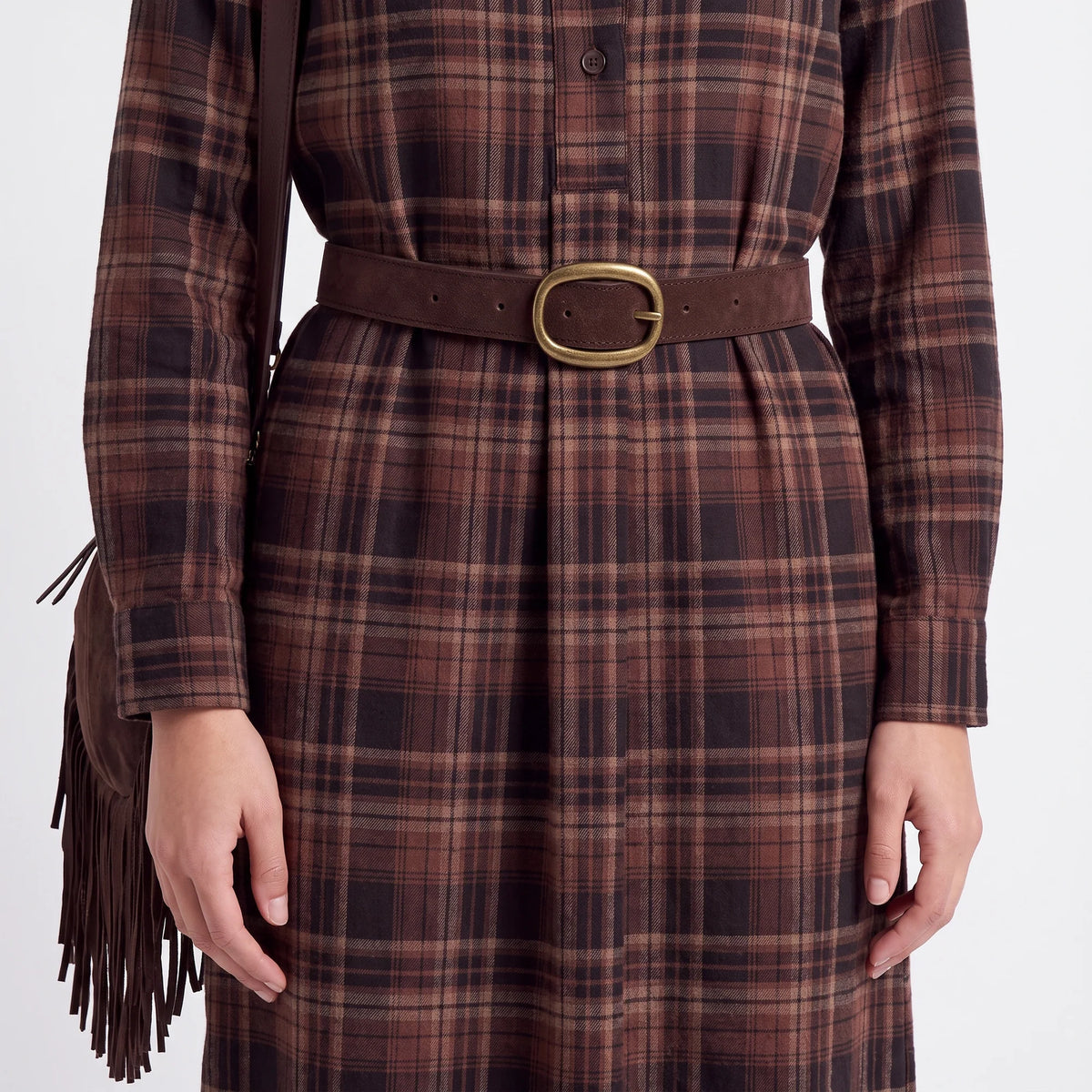 A person wearing a brown and black plaid long-sleeve dress with a matching brown belt and a gold-tone buckle, shown from the chest down against a light gray studio background.