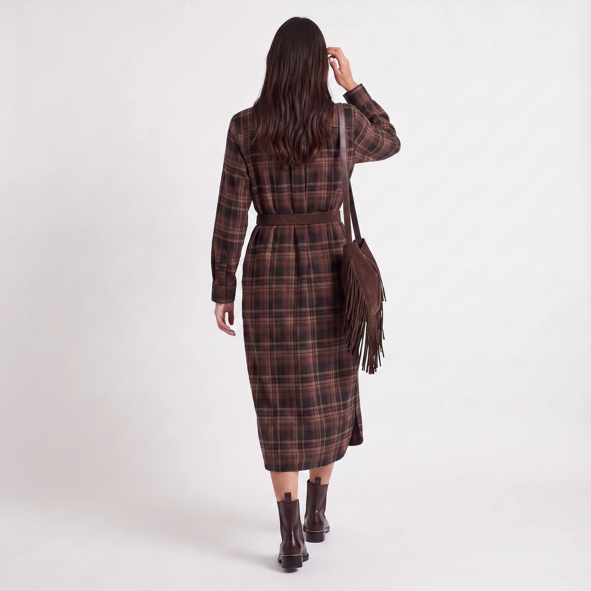 A woman is shown from behind wearing a brown and black plaid long-sleeve midi dress with a matching belt, styled with a brown fringed shoulder bag and dark brown ankle boots against a plain white studio background.