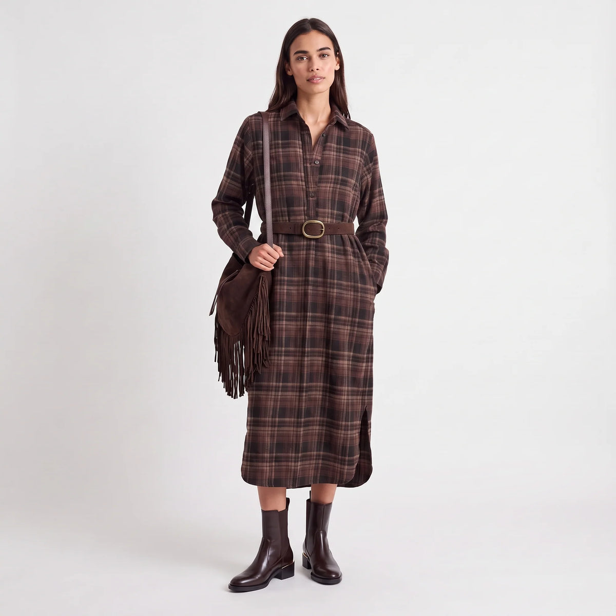 A woman stands against a light gray studio background wearing a long-sleeve brown plaid shirt dress with a matching belt, dark brown fringe bag, and dark brown ankle boots.