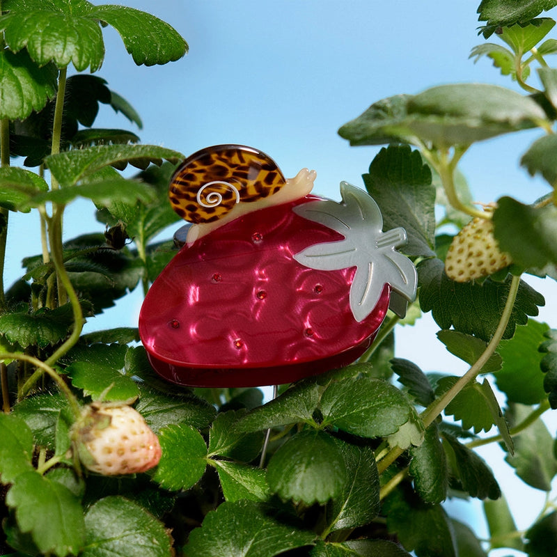 Strawberry-shaped hair clip with a snail on top, surrounded by green leaves and small strawberries.