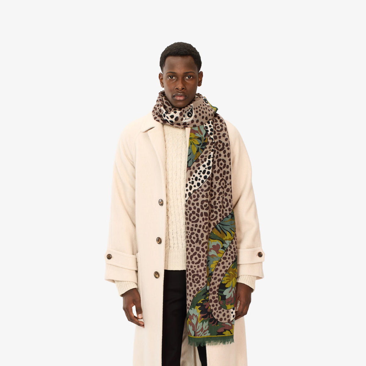 A male model wearing a cream-colored overcoat over a cream knit sweater, styled with a multi-patterned scarf featuring leopard print, green and yellow floral motifs, and black trousers, shot against a solid off-white background.
