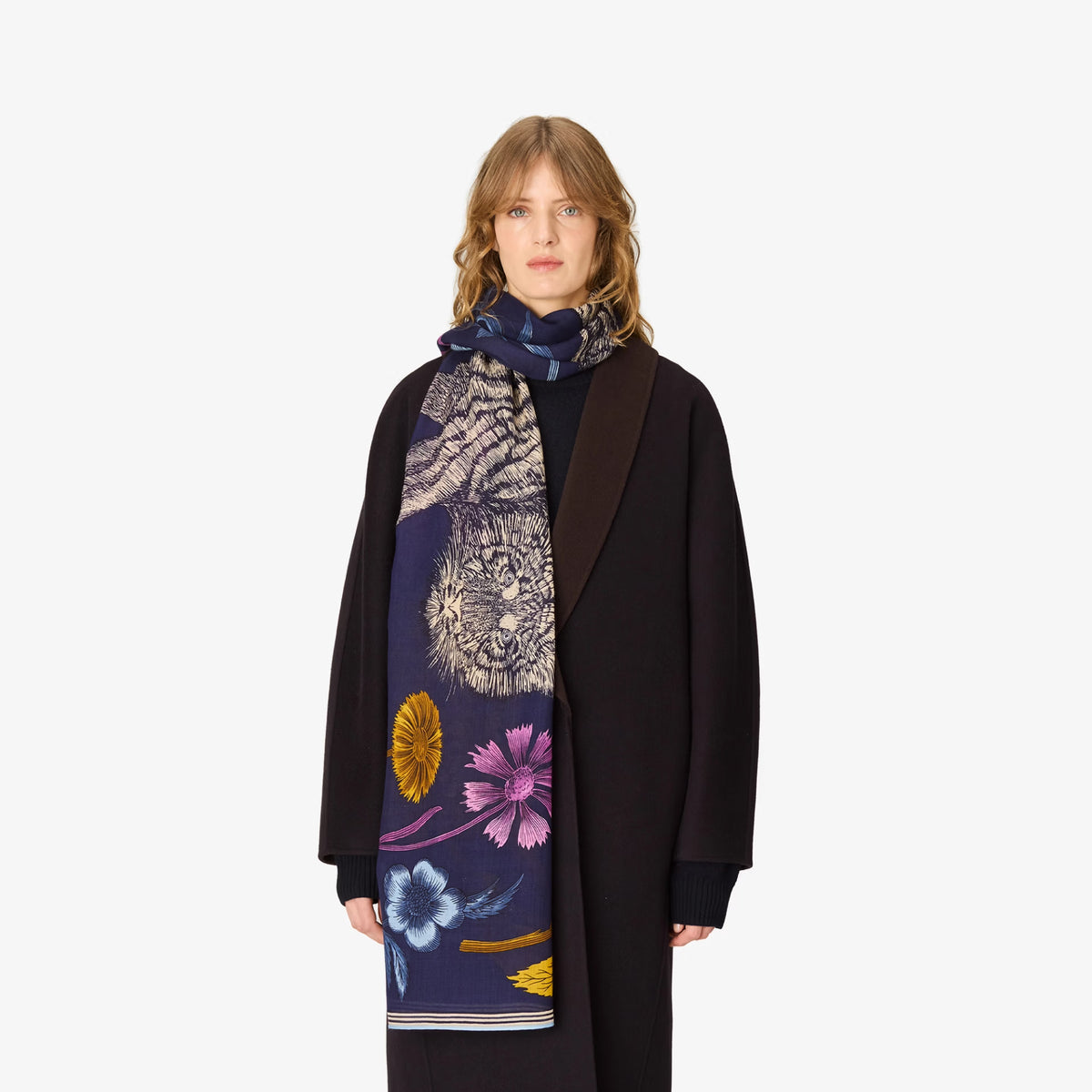 A model wearing a long black coat and a dark navy scarf with a detailed print of a tiger's face and colorful flowers, set against a plain light gray background.