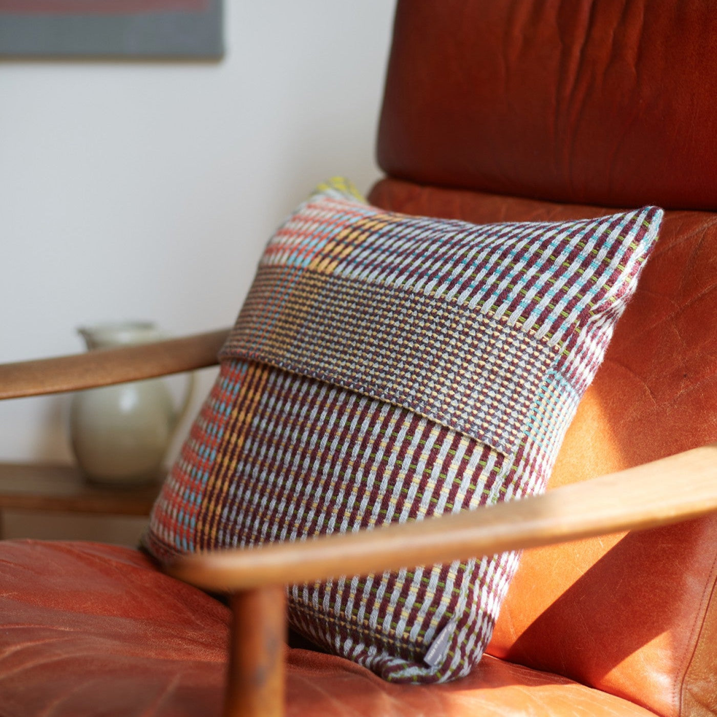 Multicolored woven throw pillow with a textured pattern in shades of maroon, beige, and teal, resting on a leather armchair.