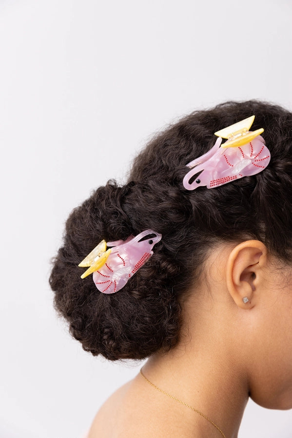 Two pink and yellow prawn shaped hair clips holding up a dark curly bun hairstyle on a model, shown from a side-back profile against a light gray background.