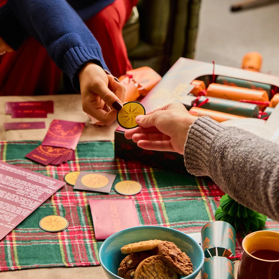A festive close-up of hands exchanging round gold tokens on a decorated table. The scene includes a plaid runner, cookies in a bowl, red envelopes, and a box of fire crackers, with two people wearing blue and gray sweaters.