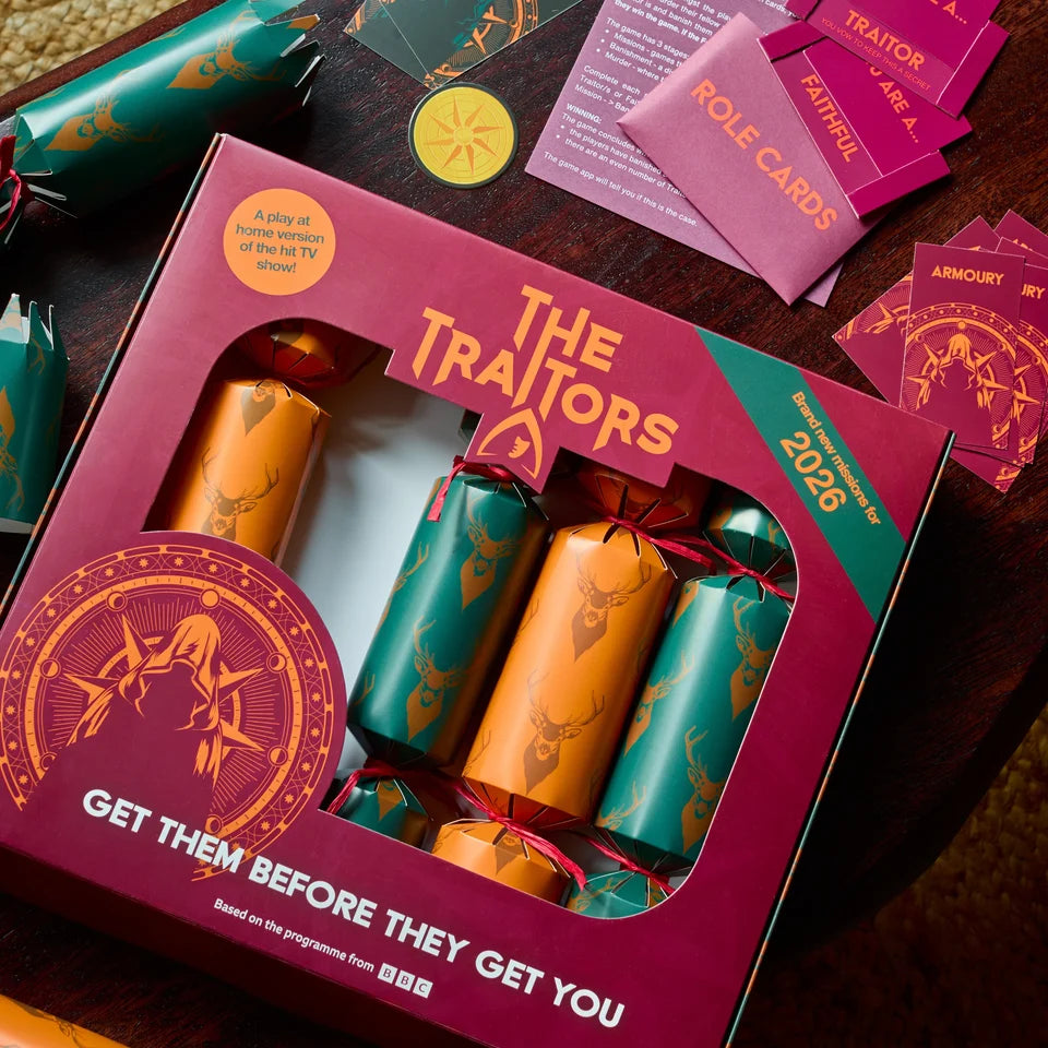 A boxed set of decorative Christmas crackers from The Traitors 2026, displayed on a dark wooden table with role cards and other themed items. The box is deep magenta with gold foil graphics and a clear window showing four crackers in teal and orange with deer designs.