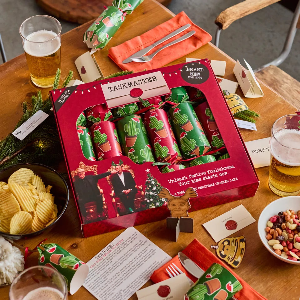 A boxed set of Taskmaster Christmas cracker cards arranged on a wooden table with snacks, drinks, and cards around it. The main product is a red gift box containing several green and red cactus-patterned cracker packs.