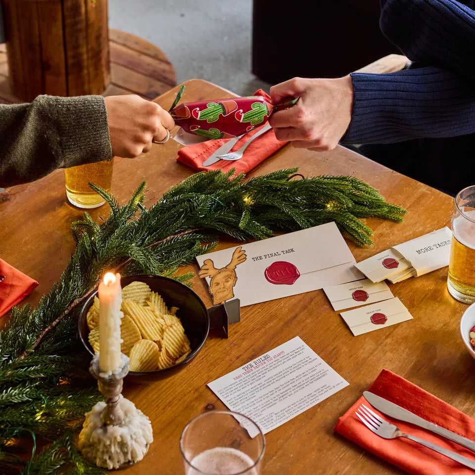 A close-up of a person's hands wrapping a gift in festive red Christmas paper with green pine branches and holiday cards on a wooden table.