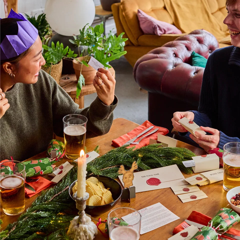 Two people sitting at a wooden table during a cozy holiday gathering, playing with cards and handling small wrapped gift boxes. The table is decorated with evergreen branches, a lit candle, glasses of beer, and festive cards and seals.