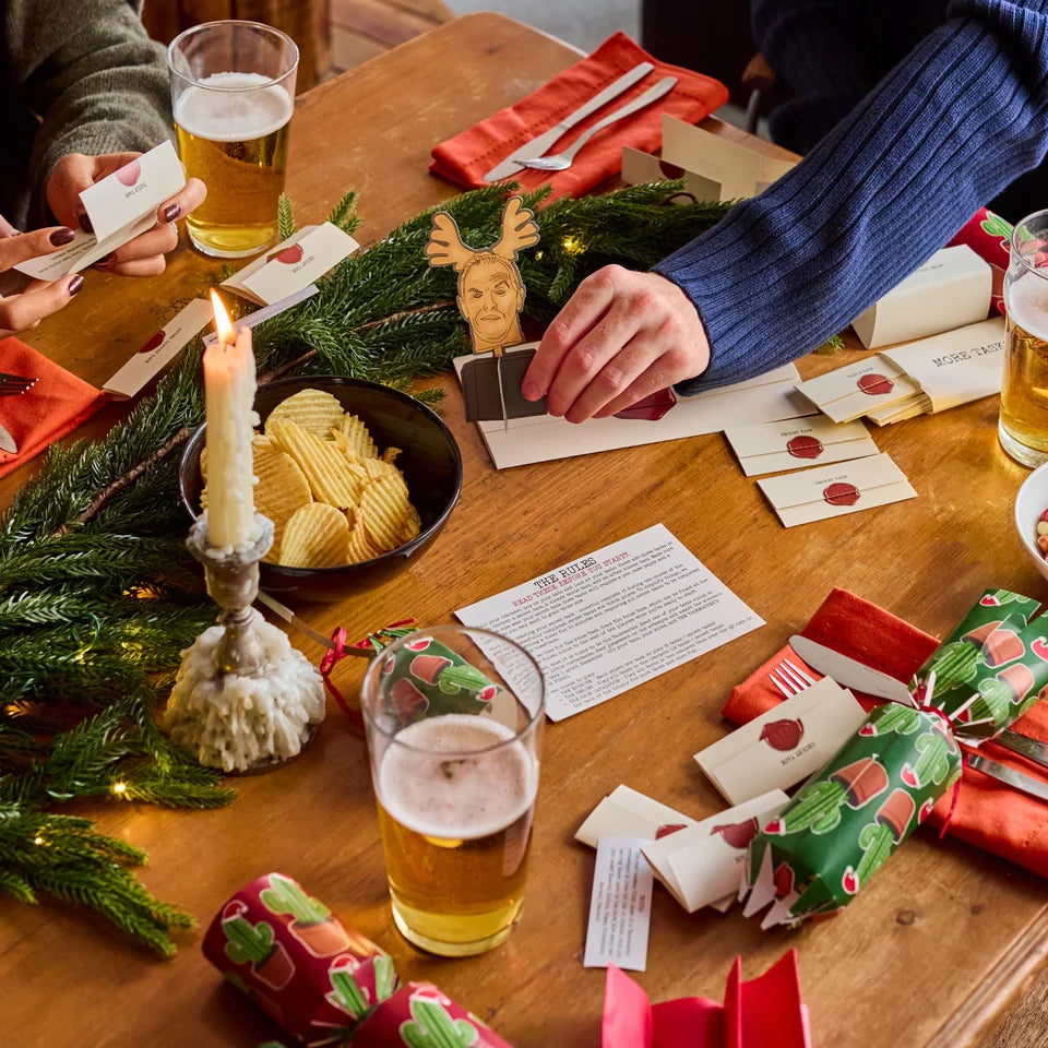 A festive tabletop scene with people placing cards and interacting with Christmas-themed items, including wrapped crackers, a candle, greenery, and printed cards on a wooden table.