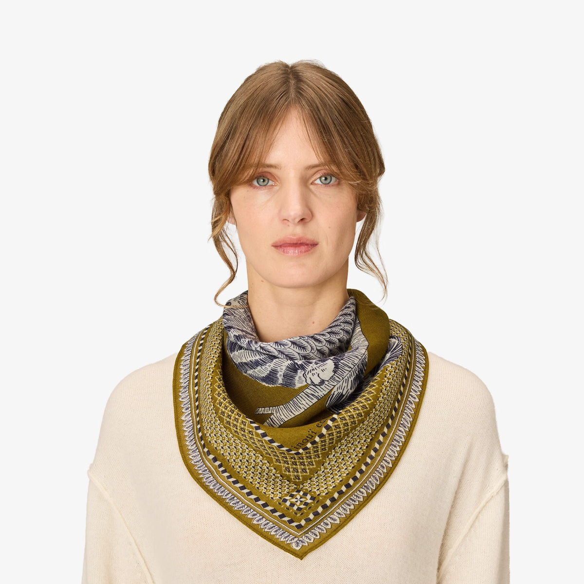 A woman wearing a cream-colored knit sweater and a patterned scarf in olive green, gray, and white, photographed against a solid light gray background.
