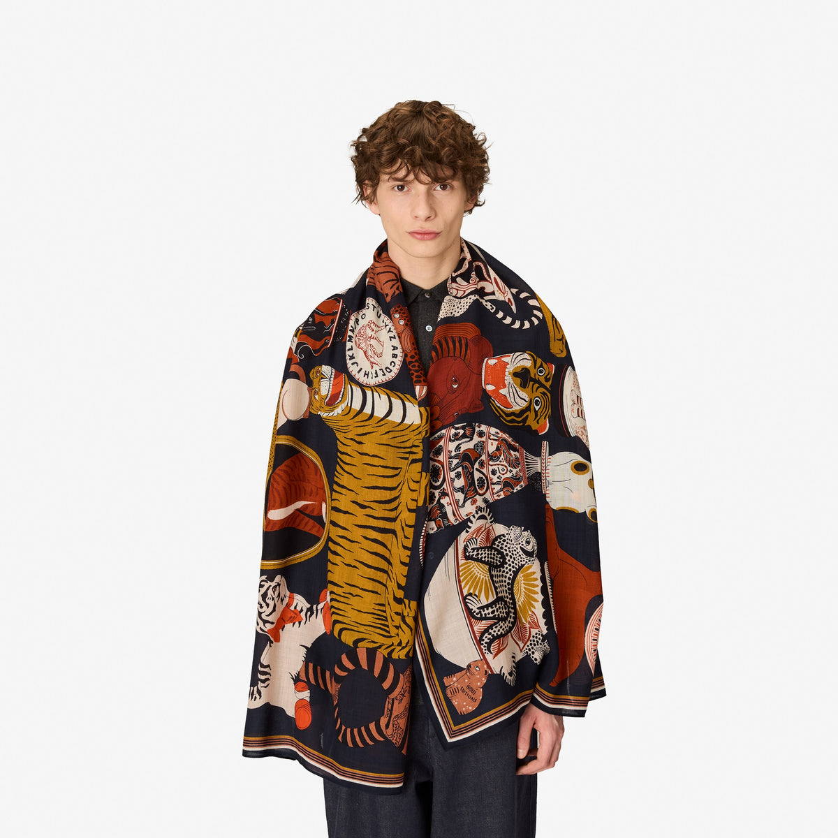 A model with curly brown hair stands against a plain white background wearing a dark navy blue silk scarf with a highly detailed, colorful tiger and animal print.