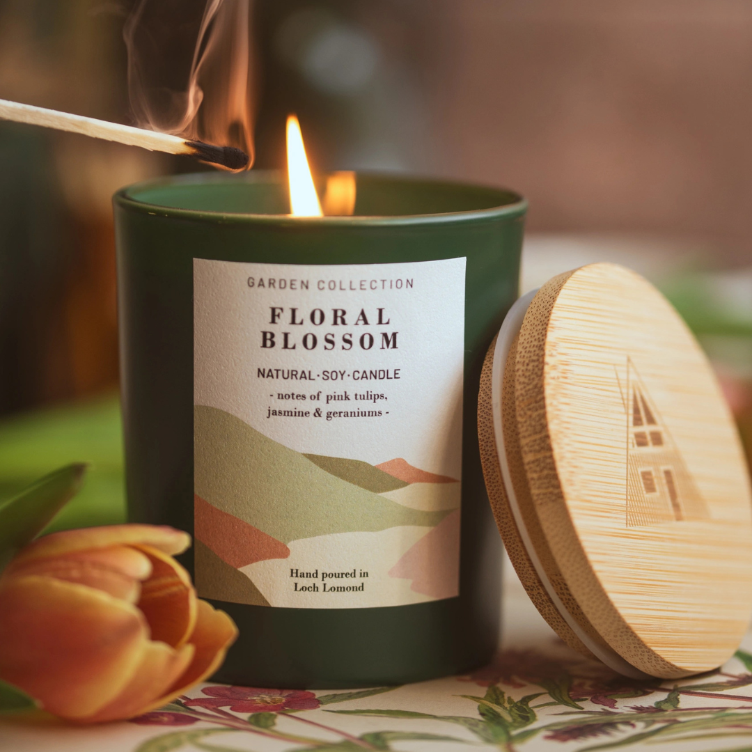 A dark green candle labelled 'Floral Blossom' by Garden Collection, lit with a match, shown with a wooden lid and a tulip flower on a floral surface.