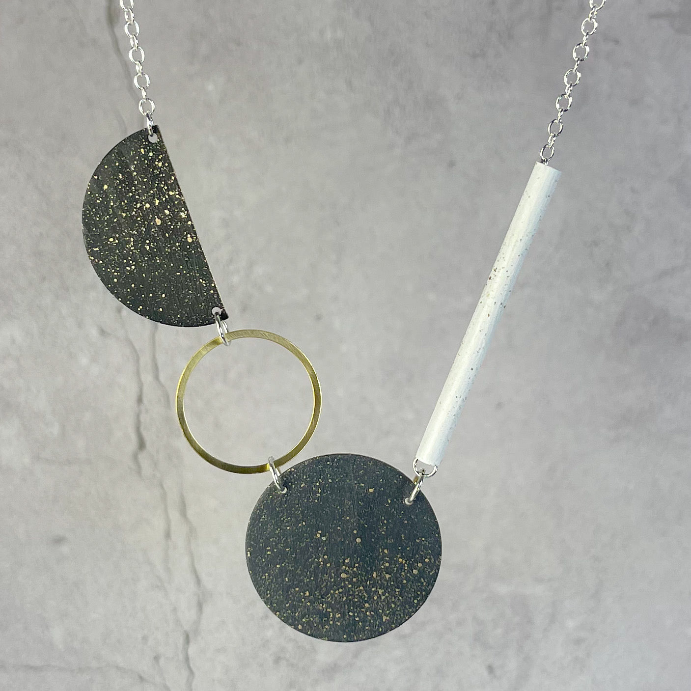 Modern necklace featuring dark speckled circular and semi-circular elements, a gold ring, and a white cylindrical pendant,