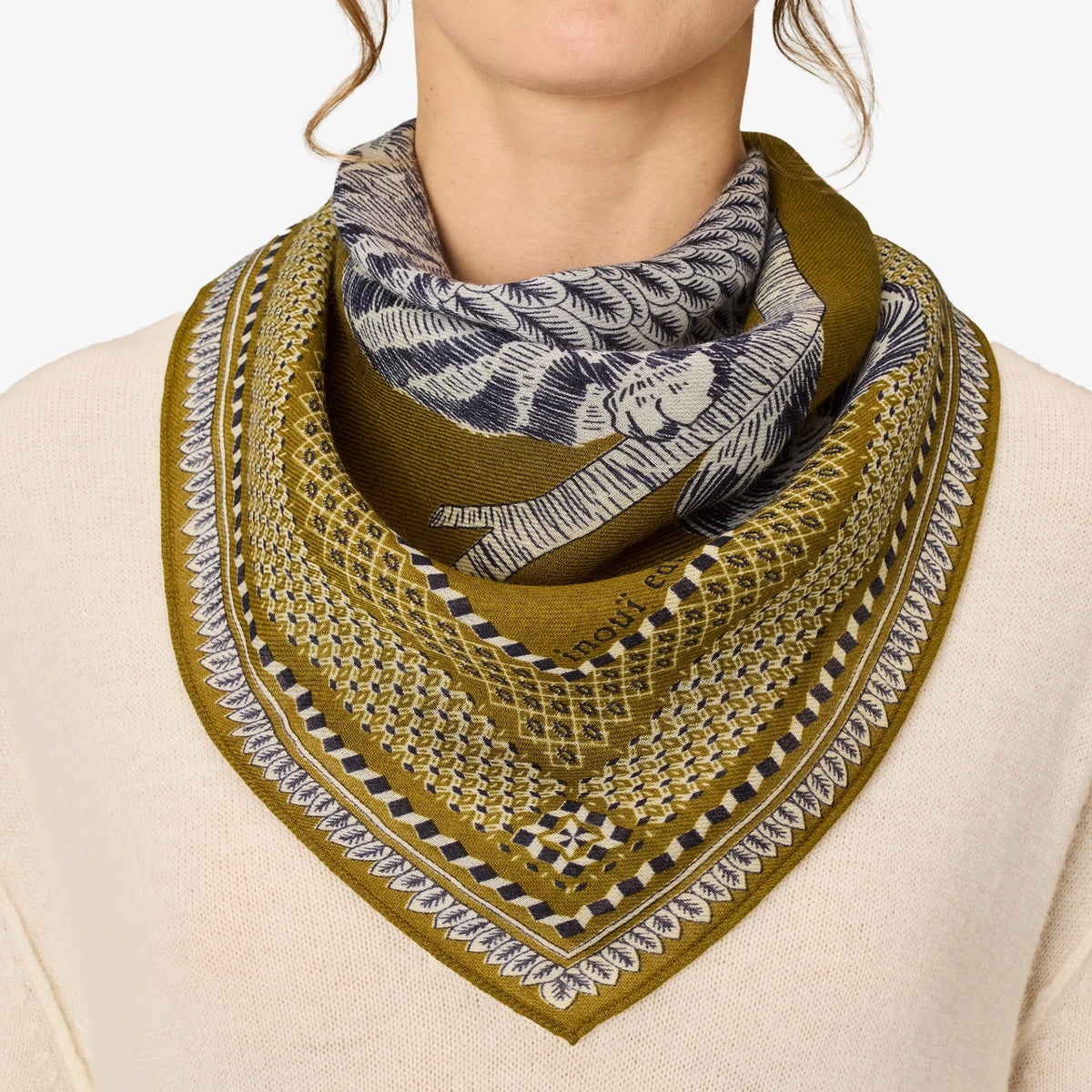 A close-up of a model wearing a square scarf styled as a bandana. The scarf features a complex pattern with an olive green border, a black and white leaf design on the top fold, and a geometric grid pattern on the bottom fold.
