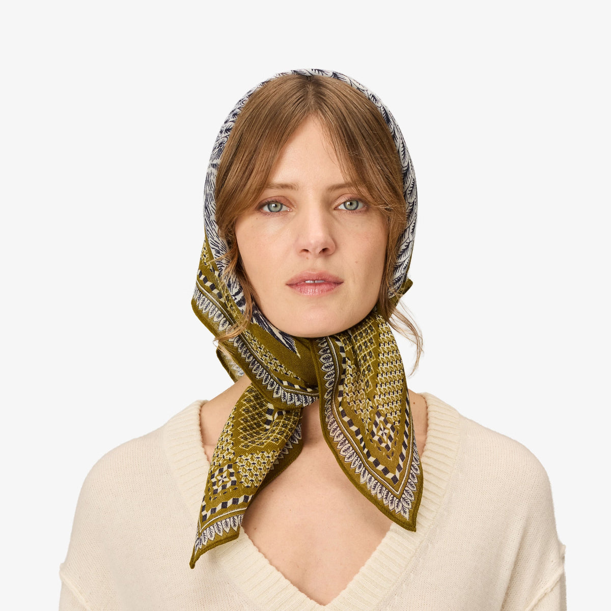 A woman wearing a patterned silk headscarf in olive green, beige, and gray, paired with a cream-colored knit sweater, set against a plain light gray background.